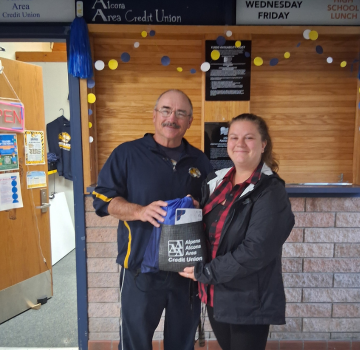 two people stanind in front of a wooden wall with blue and yellow alcona high school logos onit. A feamale is presenting a male a check for winning teacher of the month. The make is bald with glasses wearing a blue coat and tan pants holding a bag that says aaacu. The female is short with dark hair and pale skin wearing a blue aaacu shirt and jeans